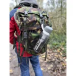 Active Pure Clear® water filter bottle attached to a camouflage backpack in a forest setting, ideal for outdoor activities and hydration.