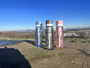 Pure Clear cycling water bottles in blue, black, and pink against a scenic outdoor backdrop, emphasising hydration for cyclists.