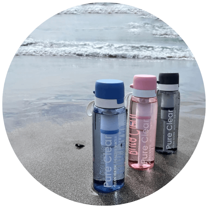 Three Pure Clear filtered water bottles in blue, pink, and grey, placed on a sandy beach with ocean waves in the background, showcasing advanced filtration technology for safe drinking water.