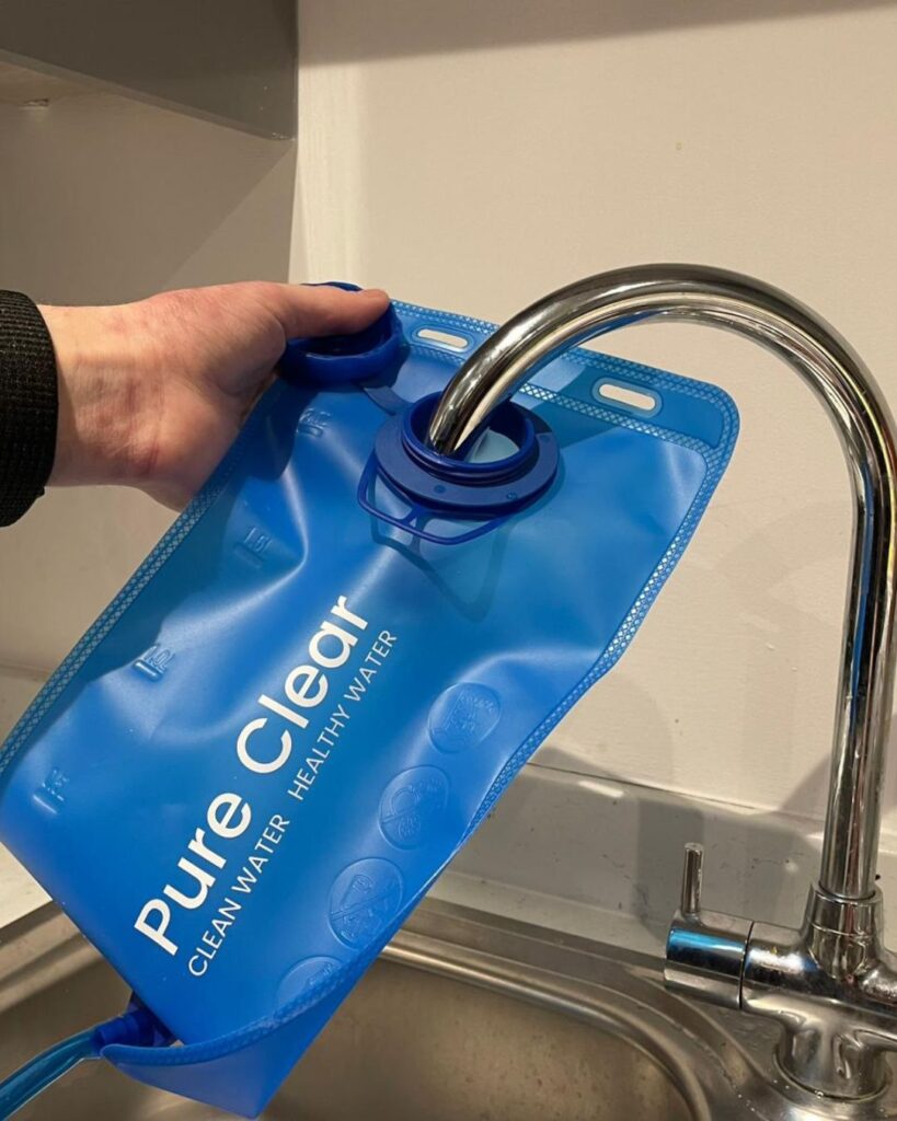 Hand holding a blue Pure Clear water bag under a tap, highlighting the brand's focus on clean and healthy water filtration solutions.