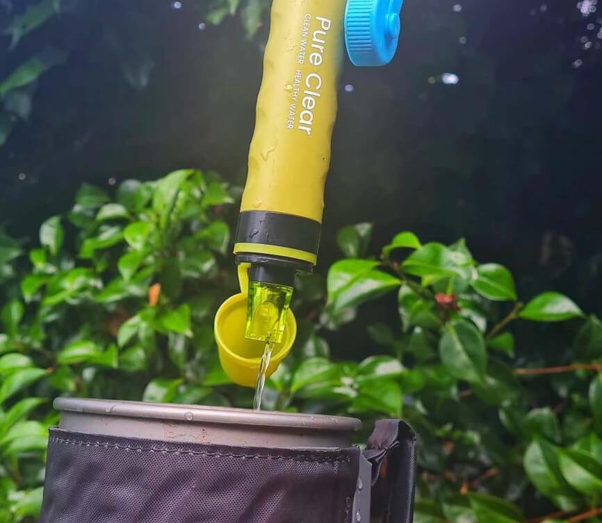 Pure Clear® water filter bottle dispensing clean water into a container, surrounded by greenery, highlighting outdoor hydration solutions.