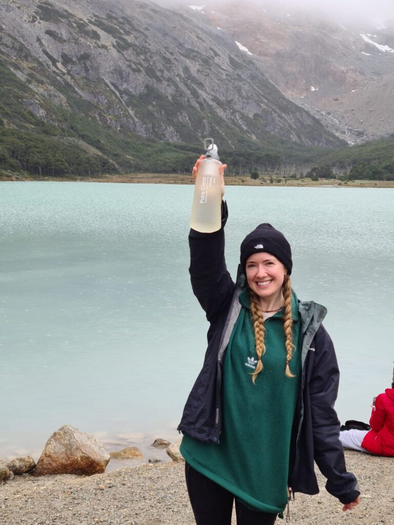 Woman holding a Pure Clear water filtration bottle above her head, standing by a turquoise lake surrounded by mountains, showcasing the product's use in outdoor settings.