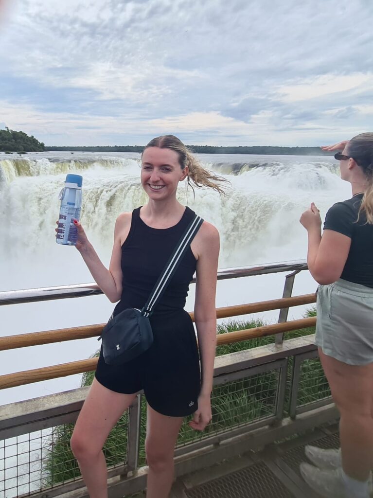 Young woman holding a Pure Clear water filter bottle in front of a waterfall, showcasing hydration solutions for outdoor activities.