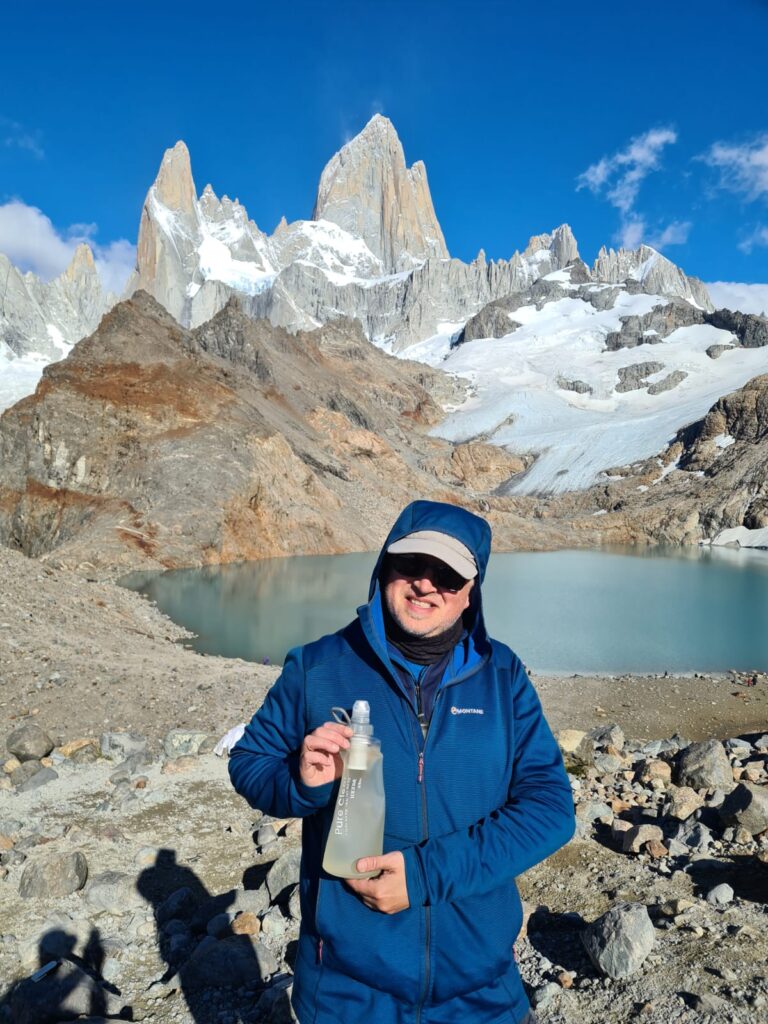 Man holding Pure Clear water filtration bottle in front of mountainous landscape with glacier and lake, showcasing outdoor hydration solution for hiking.