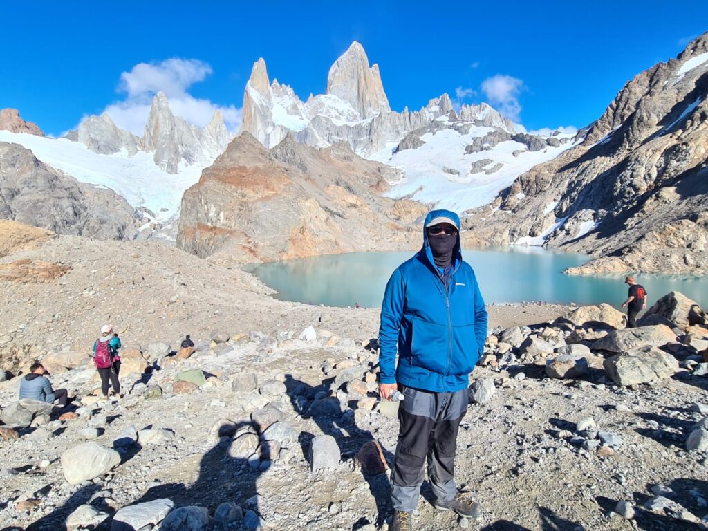 Hiker in blue jacket and mask standing on rocky terrain near turquoise glacial lake and mountains, showcasing outdoor adventure in Patagonia.