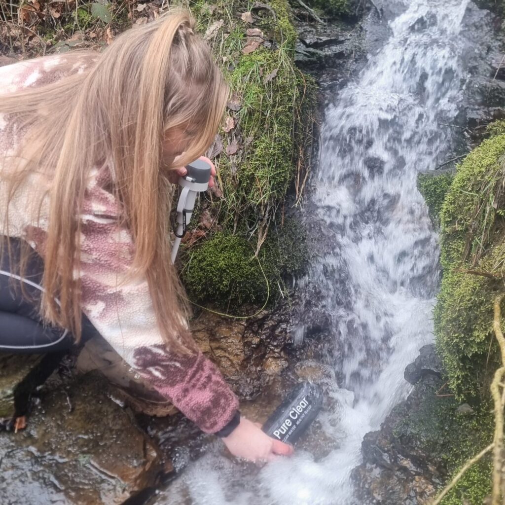 Woman using a Pure Clear® Active Water Filter Bottle to collect water from a flowing stream surrounded by mossy rocks and greenery.