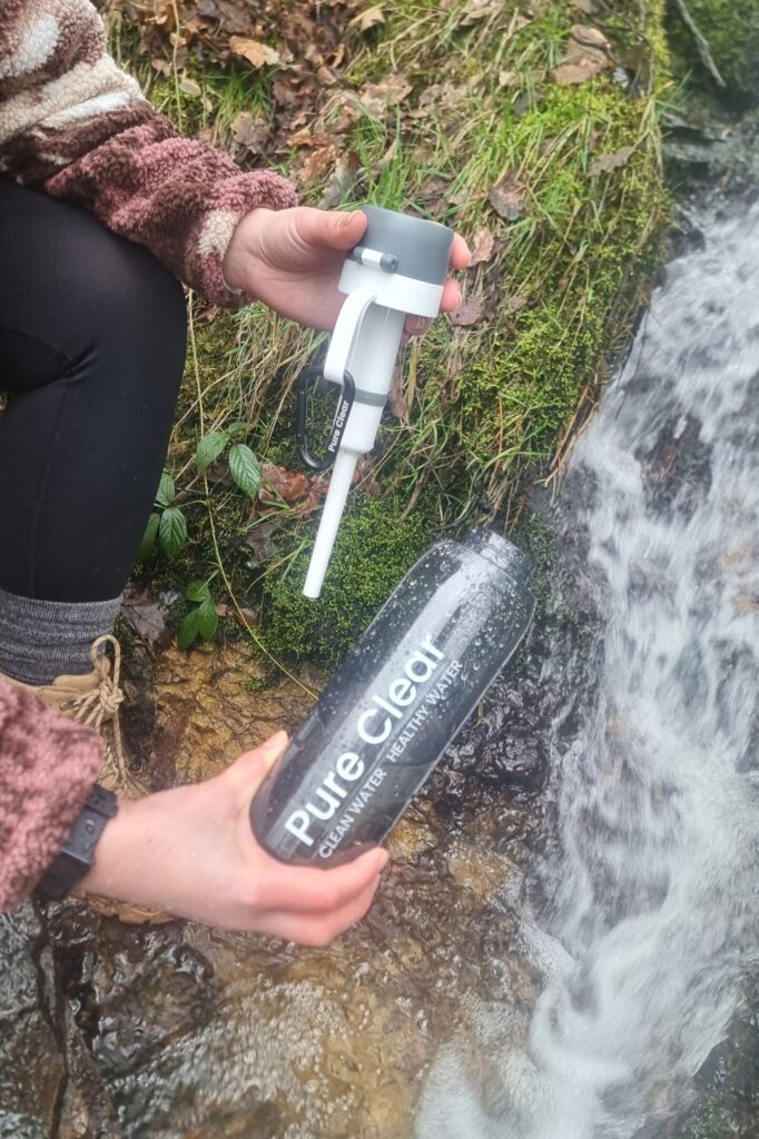 Person holding a Pure Clear water filtration bottle and filter, positioned by a flowing stream in a natural setting, showcasing outdoor hydration solutions for hikers.