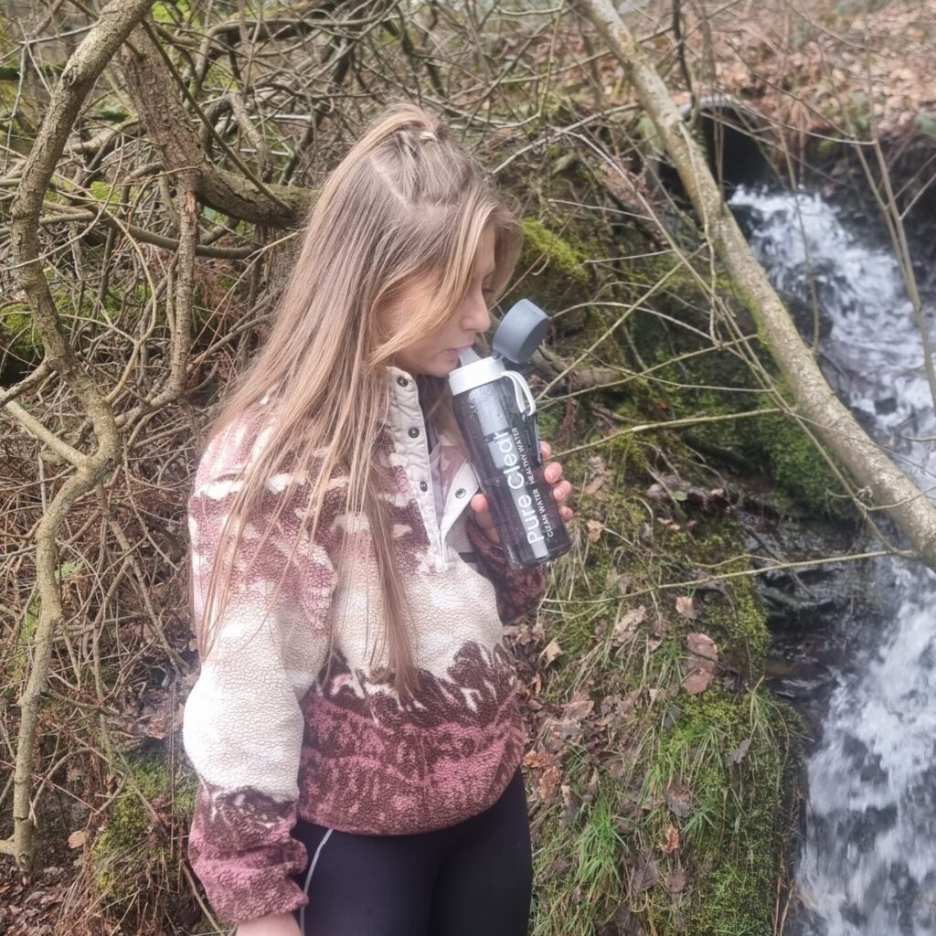 Young person drinking from a Pure Clear Filter Active Water Bottle beside a flowing stream, surrounded by lush greenery and mossy rocks, illustrating hydration and outdoor exploration.